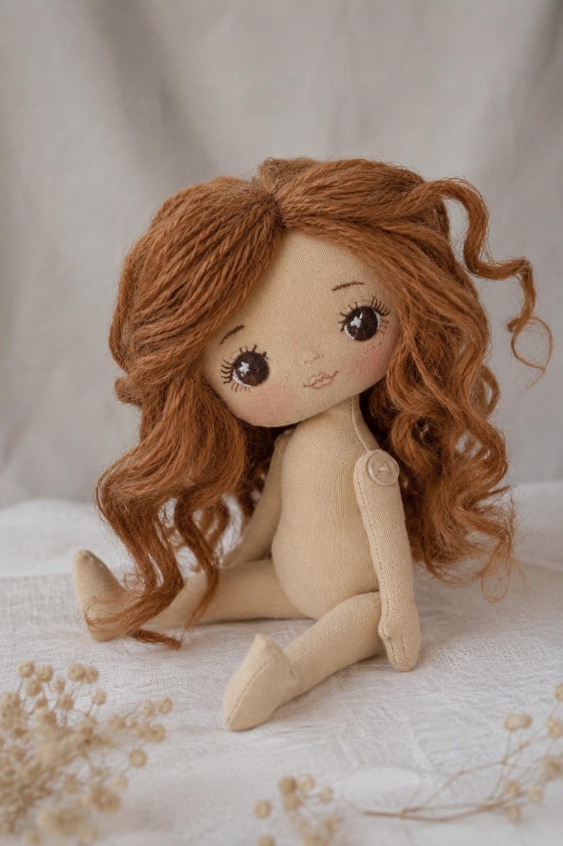 May include: A handmade fabric doll with long, wavy auburn yarn hair, dark brown embroidered eyes with long lashes, and a small stitched mouth. The doll has a light beige cloth body with a visible button joint at the shoulder and sits upright.