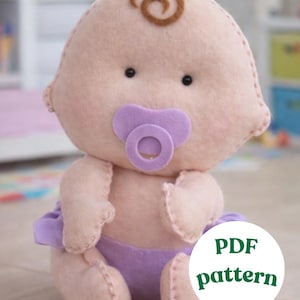 May include: A handmade felt baby doll in soft pink with a brown swirl of hair, black eyes, and a purple pacifier. The doll wears a purple diaper and is a PDF pattern.