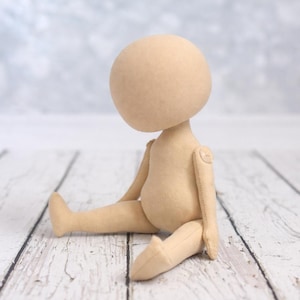 May include: A beige fabric doll with a large round head and a featureless face. The doll has articulated arms attached with visible circular joints and is posed in a seated position with legs extended forward, crafted from a smooth, solid-colored textile.
