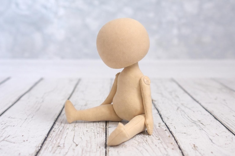 May include: A beige fabric doll with a large round head and a featureless face. The doll has articulated arms attached with visible circular joints and is posed in a seated position with legs extended forward, crafted from a smooth, solid-colored textile.