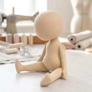 May include: A beige fabric doll with a large round head and a seated posture. The doll features simple stitched seams, a smooth surface, and articulated shoulder joints with visible circular fabric connectors on each side of the upper torso.