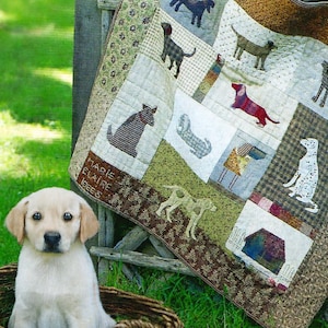 May include: A Labrador puppy in a wicker basket beside a patchwork quilt with dog illustrations. The quilt, featuring various dog breeds, is draped over a wooden fence. The scene is set outdoors on green grass.
