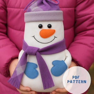 Könnte beinhalten: Ein handgemachter Schneemann-Plüsch mit lila Hut, Schal und Fäustlingen. Der Schneemann hat ein lächelndes Gesicht mit schwarzen Augen und einer orangenen Nase. Der Text "PDF PATTERN" befindet sich in einem weißen Kreis.
