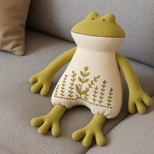 Op de afbeelding: Een pluche kussen in de vorm van een kikker met een lichtgroene kop, armen en benen en een cr&egrave;mekleurig lichaam. Het lichaam heeft geborduurde groene plantenmotieven. De kikker zit op een grijze bank naast een beige kussen.
