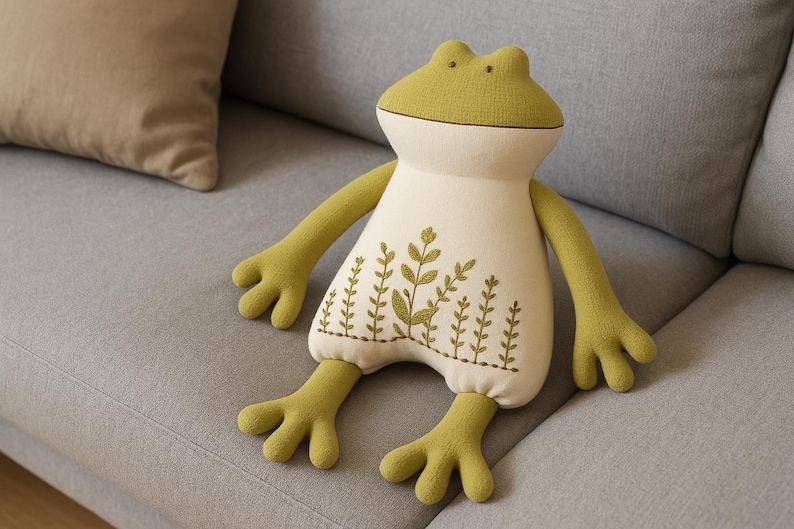 Op de afbeelding: Een pluche kussen in de vorm van een kikker met een lichtgroene kop, armen en benen en een cr&egrave;mekleurig lichaam. Het lichaam heeft geborduurde groene plantenmotieven. De kikker zit op een grijze bank naast een beige kussen.