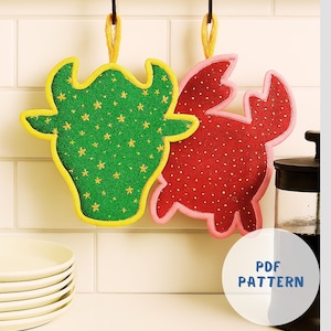 May include: Two decorative kitchen potholders shaped like a cow and a crab, hanging from a metal bar. The cow potholder is green with yellow trim and gold star accents. The crab potholder is red with pink trim and gold dot accents. A white circle with "PDF PATTERN" is in the image.