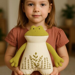 Op de afbeelding: Een pluche kikkerpop met een lichtgroene kop en ledematen en een cr&egrave;mekleurig lichaam. Het lichaam is geborduurd met een botanisch ontwerp in groentinten. De pop is zacht en knuffelig, voor kinderen.