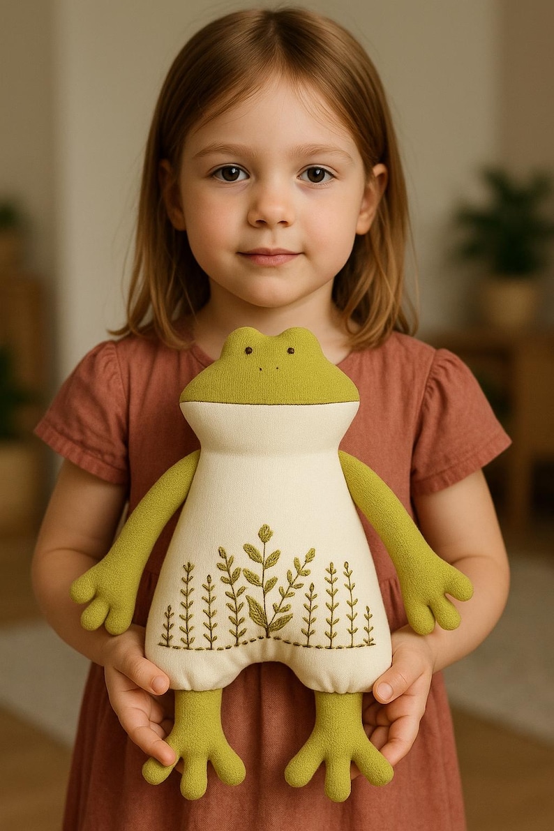 Op de afbeelding: Een pluche kikkerpop met een lichtgroene kop en ledematen en een cr&egrave;mekleurig lichaam. Het lichaam is geborduurd met een botanisch ontwerp in groentinten. De pop is zacht en knuffelig, voor kinderen.