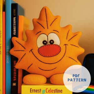 Può includere: Un peluche arancione a forma di sole sorridente con naso rosso e occhi bianchi, appoggiato su una pila di libri. I libri sono intitolati "Ernest & Celestine" e "True Heroes". Un cerchio bianco con "PDF PATTERN" è nell'immagine.