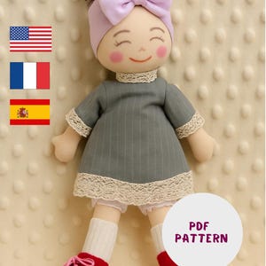 Puede incluir: Una muñeca de trapo hecha a mano con un vestido gris, ribete de encaje blanco y zapatos rojos. La muñeca tiene una diadema de lazo rosa, mejillas sonrosadas y una cara sonriente. El patrón es un PDF.