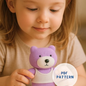 May include: A knitted purple teddy bear wearing a white tank top. The bear has a white muzzle and a black button nose. A circular graphic with the text "PDF PATTERN" is visible. The bear is held by a child.