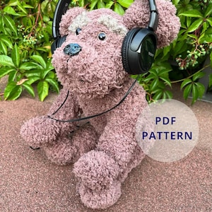 May include: A plush, mauve teddy bear wearing black Sony headphones. The bear has a light gray patch on its head and a blue nose. The image includes the text "PDF PATTERN" in a white circle.