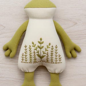 Op de afbeelding: Een pluche kikker met een lichtgroene kop, armen en benen en een cr&egrave;mekleurig lichaam. Het lichaam heeft een geborduurd ontwerp van groene planten. De kikker heeft een vriendelijke, grillige uitstraling, ideaal voor een kinderkamer.