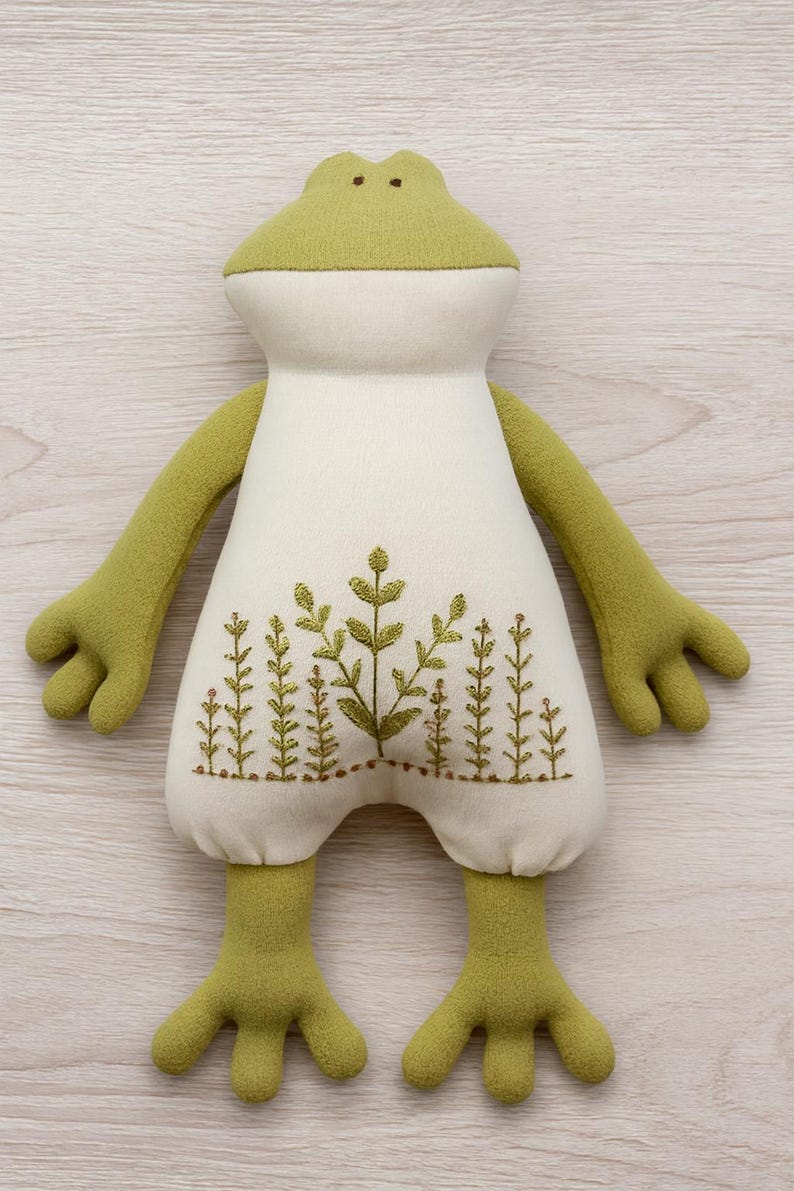 Op de afbeelding: Een pluche kikker met een lichtgroene kop, armen en benen en een cr&egrave;mekleurig lichaam. Het lichaam heeft een geborduurd ontwerp van groene planten. De kikker heeft een vriendelijke, grillige uitstraling, ideaal voor een kinderkamer.