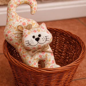 Puede incluir: Un gato de tela hecho a mano con un estampado floral en una cesta de mimbre marrón. El gato tiene una cara blanca con detalles bordados y un lazo de yute. La cesta es redonda y tejida.