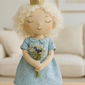 May include: A handcrafted doll wearing a light blue dress and a gold crown. The doll has closed eyes and holds a bouquet of white and blue flowers. The doll's hair is curly and blonde. The dress features a floral pattern.