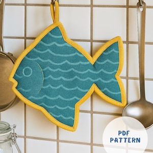 May include: A teal fish-shaped pot holder with a yellow border and hanging loop. The pot holder features a wave pattern and a stitched eye. The words "PDF PATTERN" are in a white circle. Kitchen decor.