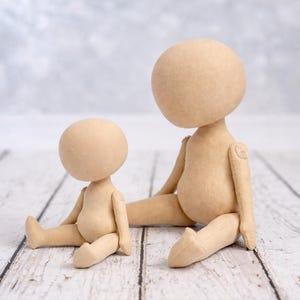 May include: Two beige fabric dolls with round, featureless heads and articulated limbs. The smaller doll is seated upright, while the larger doll is seated with one leg extended. Both dolls have a smooth, plain surface and neutral tan color.