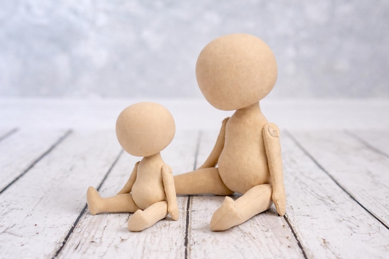 May include: Two beige fabric dolls with round, featureless heads and articulated limbs. The smaller doll is seated upright, while the larger doll is seated with one leg extended. Both dolls have a smooth, plain surface and neutral tan color.