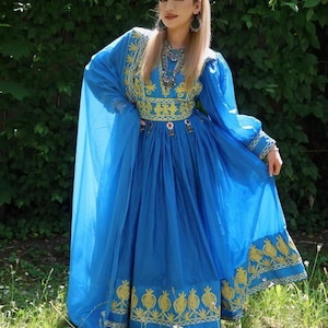 Può includere: Un abito blu acceso con maniche lunghe e gonna ampia. L'abito presenta ricami dorati intricati lungo lo scollo, le maniche e l'orlo. Uno scialle blu abbinato è drappeggiato sulle spalle. Il vestito è indossato con gioielli e un copricapo ornati.