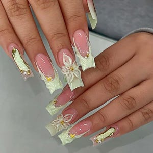 Könnte beinhalten: Lange, quadratische Acrylnägel mit hellrosa Basis und weißen Spitzen. Die Nägel weisen 3D-Blumendesigns, goldene Akzente und einen strukturierten, hellgrünen Rand auf. Einige Nägel haben goldene und perlenartige Verzierungen.