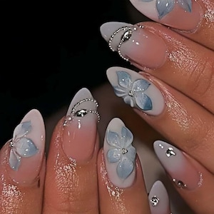 Puede incluir: Primer plano de uñas manicuradas con un degradado rosa claro y blanco. Cada uña presenta un diseño de flor azul claro en 3D con detalles plateados. Algunas uñas tienen adornos de cuentas plateadas.