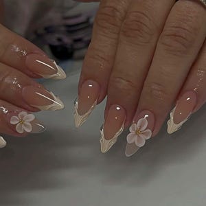 May include: A close-up of manicured nails with a nude base and white, textured accents. Some nails feature a 3D white flower with a gold center. The nails are long and pointed, with a glossy finish.