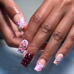 Puede incluir: Primer plano de una mano con uñas acrílicas largas y cuadradas. Las uñas presentan varios diseños, incluyendo una flor rosa 3D con una gema azul, una uña burdeos con detalles de burbujas y uñas con patrones rosas y blancos. Las uñas tienen un acabado brillante.