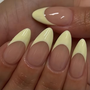May include: Close-up of manicured nails featuring a French manicure design. The almond-shaped nails have a pale yellow tip and a nude base. The nails are glossy, presenting a classic and elegant style, perfect for any occasion.