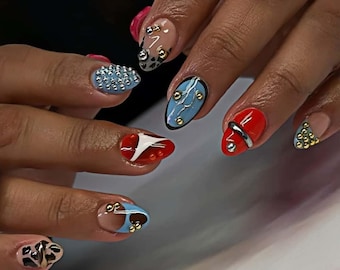 May include: Close-up of a hand showcasing various nail art designs. The nails are painted in shades of blue, red, and nude, adorned with silver and gold studs. Some nails feature abstract patterns, while others have a textured, studded finish.