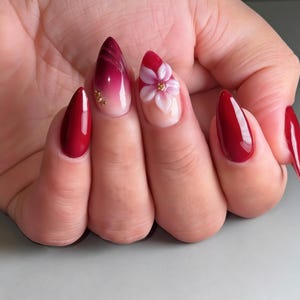 May include: Almond-shaped nails featuring a glossy deep red finish. One nail displays a dark red to light pink ombre gradient with small gold metallic studs, while another features a raised, three-dimensional light pink flower with a gold centre.