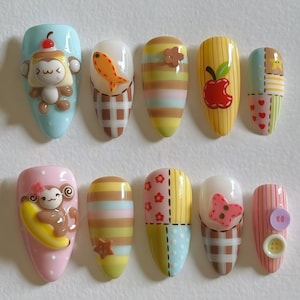 Puede incluir: Una colección de diez uñas artificiales con varios diseños. Las uñas presentan personajes de dibujos animados, frutas y patrones como rayas, patchwork y cuadros. Los colores incluyen rosa, azul, amarillo, marrón y blanco. Algunas uñas tienen adornos 3D.