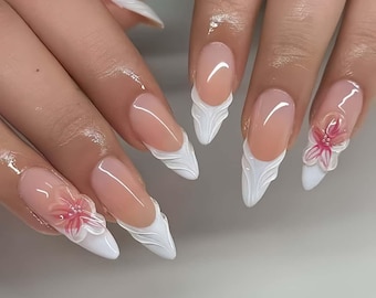 May include: Close-up of a set of long, almond-shaped acrylic nails. The nails feature a French tip design with a white, sculpted edge. Some nails have a 3D pink flower accent, adding a touch of elegance to the manicure.