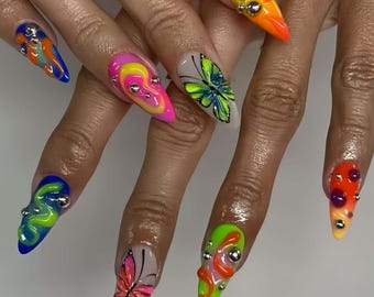 May include: A set of long, pointed acrylic nails with vibrant designs. Each nail features a unique combination of colors, including neon green, pink, orange, and blue, with abstract patterns and metallic embellishments. Some nails showcase butterfly and swirl designs.