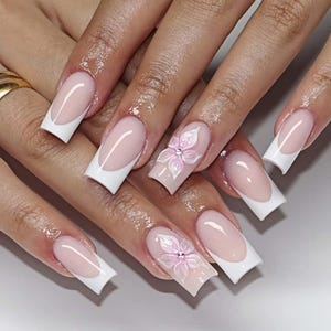 Könnte beinhalten: Ein Satz langer, quadratischer Acrylnägel. Die Nägel haben ein klassisches French-Maniküre-Design mit weißen Spitzen und einer blassrosa Basis. Zwei Nägel sind mit 3D-Blumenmustern in Rosa mit Strass-Mittelpunkten verziert.