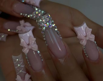 May include: Long, light pink artificial nails with a French tip design. Some nails feature 3D pink bows, while others are adorned with sparkling rhinestones. The nails are a fashionable accessory.