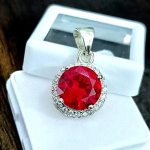 May include: A round, red gemstone pendant with a halo of small, clear stones. The pendant is set in a silver-colored metal and is displayed in a white jewelry box. The gemstone is faceted, reflecting light.