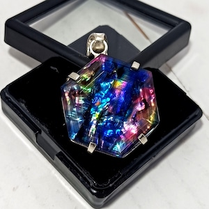 Op de afbeelding: Een hanger van dichroisch glas met een zilveren balein en accenten. De hanger heeft een veelzijdige zeshoekige vorm en toont een levendig kleurenpalet, waaronder blauw, paars, roze en groen. Het ligt op een zwarte sieradendoos.