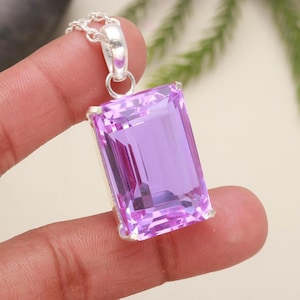 May include: A rectangular, light purple gemstone pendant with a silver chain. The gemstone is faceted, reflecting light, and set in a silver bezel. The pendant is held by a hand against a soft background.