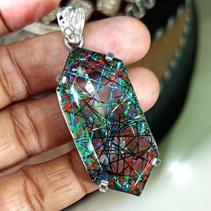 May include: A rectangular pendant with a clear, faceted stone featuring colourful, intersecting lines of red, green, blue, and black. The pendant is held by a silver-coloured bail and four silver-coloured prongs.