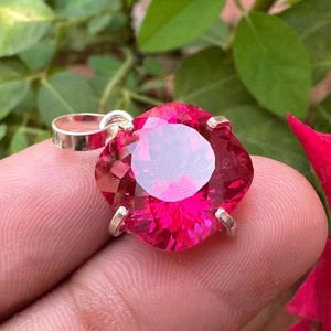 May include: A vibrant, faceted, hot pink gemstone pendant with a silver bail. The gemstone is a large, round shape with a flower-like setting. The pendant is displayed against a background of green leaves and a red flower.