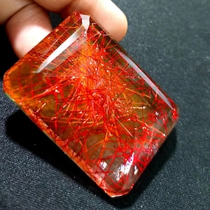 以下が含まれることがあります： 鮮やかな赤いインクルージョンがネットワーク状になった、長方形の透明な宝石。 宝石は研磨された表面を持ち、暖かく琥珀色のオレンジ色をしています。 赤い線が印象的な視覚的パターンを作り出しています。