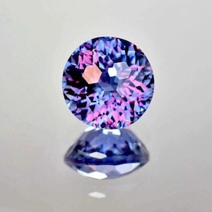 Op de afbeelding: Een ronde, gefacetteerde edelsteen met een mix van blauwe, paarse en roze tinten. De edelsteen wordt weerspiegeld op een glad oppervlak, waarbij de symmetrische slijpvorm en kleurvariaties worden benadrukt. De edelsteen lijkt te zweven.