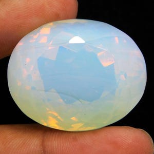 Puede incluir: Una gran piedra preciosa de ópalo facetada de forma redonda. El ópalo muestra un color base blanco lechoso con sutiles destellos de amarillo y azul. La gema se sostiene entre dos dedos sobre un fondo negro.
