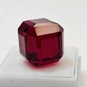 Może przedstawiać: Fasetowany, prostokątny, ciemnoczerwony kamień szlachetny. Klejnot jest prezentowany na białej powierzchni, ukazując jego przejrzystość i bogaty kolor. Szlif kamienia tworzy wiele odbijających światło powierzchni.
