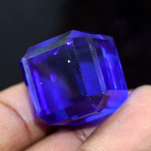 May include: A square-cut, deep blue gemstone. The gem is held in a hand against a dark background, highlighting its facets and colour. The facets reflect light, creating a sparkling effect. The gem is a vibrant shade of blue.