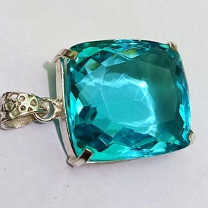 Op de afbeelding: Vierkante, lichtblauwe edelsteen hanger met een zilveren zetting. De edelsteen heeft een levendige turkooizen kleur en een facetgeslepen oppervlak. De hanger heeft een decoratieve balein om aan een ketting te bevestigen. Het ontwerp is elegant en opvallend.