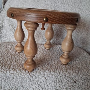 Könnte beinhalten: Ein kleiner Holzhocker mit einer runden Sitzfläche, die eine sichtbare Holzmaserung und messingfarbene Metallnieten am Rand aufweist. Der Hocker steht auf vier hellen, gedrechselten Holzbeinen mit einer glatten Oberfläche und bauchiger Form.