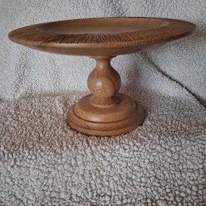 May include: A light brown wooden pedestal bowl featuring a wide, shallow circular top with a textured, grooved rim and a smooth, rounded central stem supported by a sturdy, tiered circular base with a visible natural wood grain pattern.
