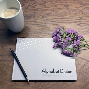 Könnte beinhalten: Ein Buch mit dem Titel "Alphabet Dating" und dem Untertitel "52 Dateideen - eine Liebesreise von A bis Z" liegt auf einer Holzoberfläche. Ein schwarzer Stift liegt auf dem Buch, daneben ein Zweig mit lila Blumen und eine weiße Tasse mit Kaffee.